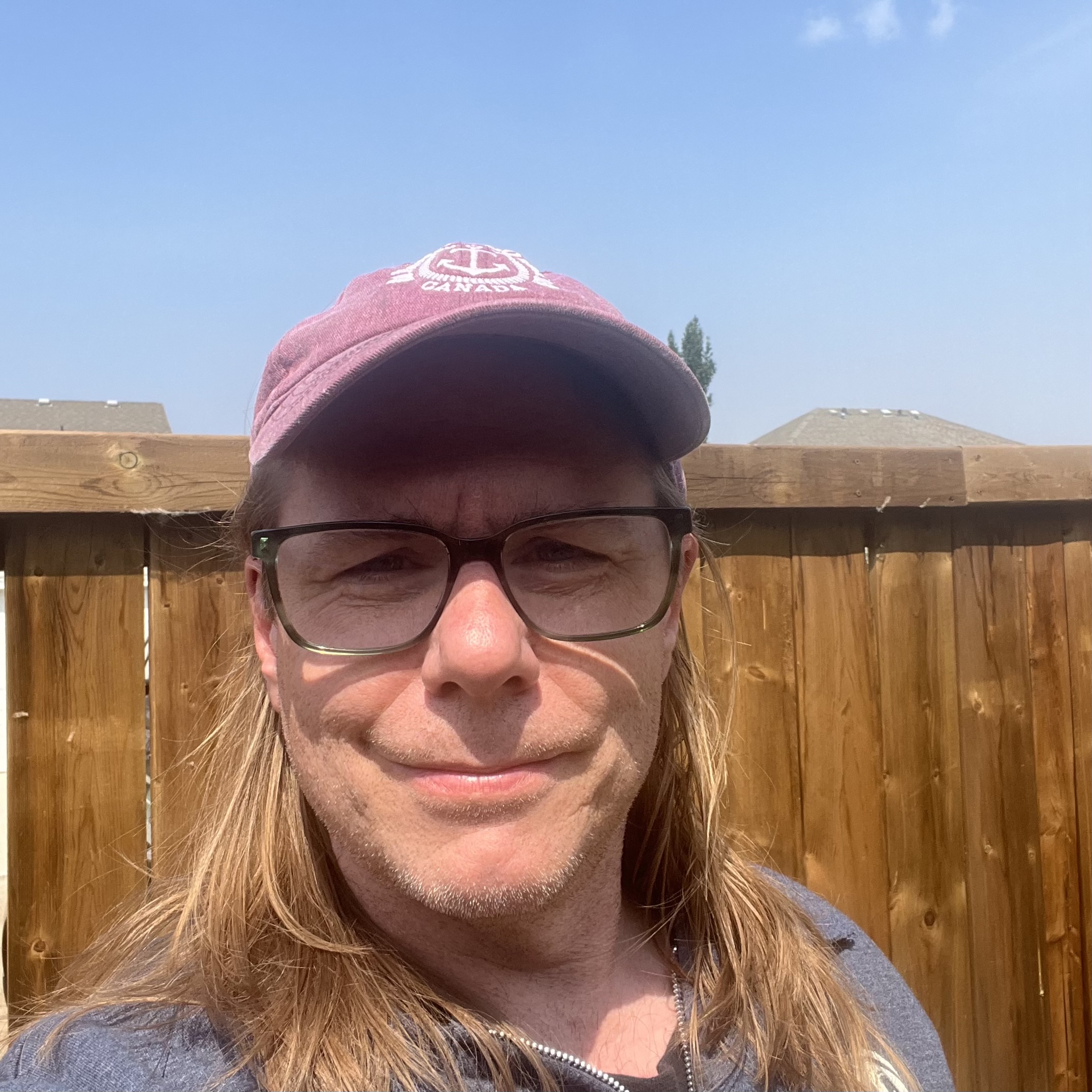 Councillor Ryan Pollard, June 2025. Sunny day, a fence in the background, he has long hair and is wearing a baseball cap and hoodie